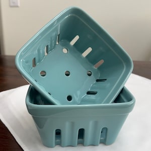 May include: Two square, light blue ceramic berry baskets with a glossy finish. Each basket features a series of vertical rectangular slots along the sides and circular drainage holes on the bottom, designed to mimic the look of traditional produce containers.