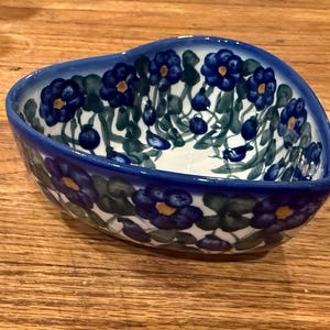 May include: Heart-shaped ceramic bowl with a blue floral pattern. The bowl has a dark blue rim and is decorated with blue flowers and green leaves. The interior is white. The bowl is sitting on a wooden surface.