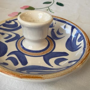 May include: A ceramic candle holder with a central cup and a wide, shallow dish. The dish is decorated with blue abstract patterns on a white background, with a brown rim. The candle holder is on a white tablecloth.