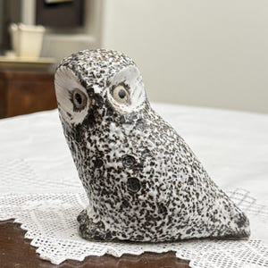 May include: A ceramic owl figurine featuring a textured, speckled black and white glaze. The owl has large, round, light-coloured eyes set within white facial discs, a pointed beak, and a compact, rounded body shape with a mottled surface finish.