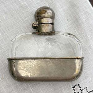 May include: A vintage, clear glass flask with a rounded top and a silver-coloured metal base and cap. The flask has a rectangular shape with rounded edges. The metal components show signs of age and wear.