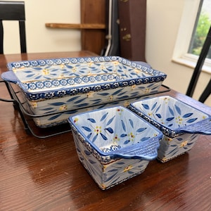 May include: A rectangular ceramic baking dish with a blue and yellow floral pattern, resting in a black metal wire stand, accompanied by two matching square ceramic bowls with blue handles, all featuring a consistent hand-painted design.