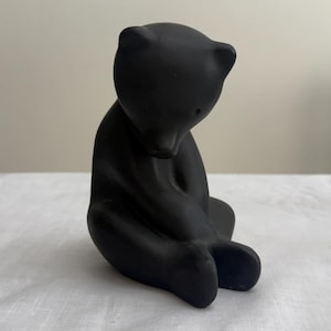 May include: A matte black figurine of a bear in a seated, hunched position with its head bowed downward. The bear has rounded ears, a smooth surface, and a solid, opaque appearance, captured from a slightly elevated side angle view.
