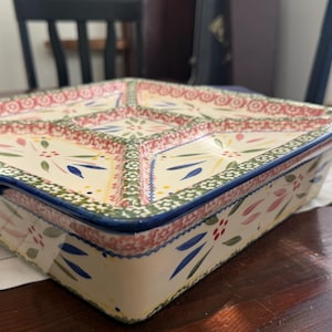 May include: A square, divided serving dish with a cream-colored base. The dish features a floral pattern in red, green, and blue, with a dark blue rim and handles. It is placed on a white, patterned tablecloth.