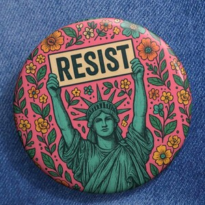 Op de afbeelding: Een roze knoop met een bloemenontwerp met een groen beeld van het Vrijheidsbeeld dat een bord vasthoudt met de tekst "RESIST".