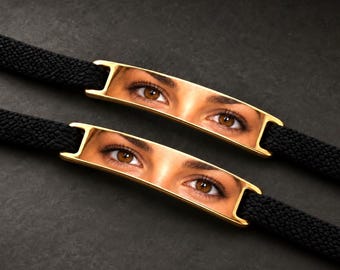 May include: Two bracelets featuring black woven fabric bands and gold-toned metal rectangular plates. Each plate displays a high-resolution, realistic print of a pair of brown eyes, capturing detailed irises, eyelashes, and skin texture.