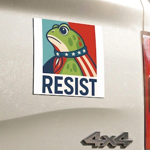 May include: A rectangular bumper sticker featuring a green frog with a determined expression, wearing a blue collar with stars and a red, white, and blue cape. The word "RESIST" is printed in bold white letters at the bottom.