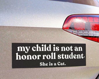 May include: A rectangular black bumper sticker with white text that reads, my child is not an honor roll student. She is a Cat. The sticker is applied to the rear of a silver vehicle, positioned below the rear window and above the bumper.