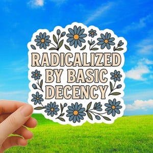 Op de afbeelding: Een sticker met de tekst "RADICALIZED BY BASIC DECENCY" omringd door blauwe bloemen en groene bladeren. De sticker wordt vastgehouden tegen een achtergrond van een helderblauwe lucht en groen gras.