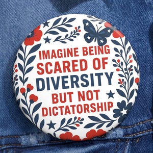 Op de afbeelding: Een ronde button met een witte achtergrond, met de tekst "IMAGINE BEING SCARED OF DIVERSITY BUT NOT DICTATORSHIP" in rood en blauw. De button is versierd met bloemen- en vlindermotieven in rood en blauw.