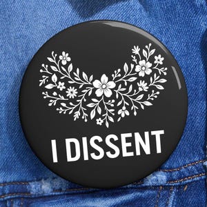May include: A black badge with the white text "I DISSENT" below a white floral design. The round badge is set against a blue denim jacket.