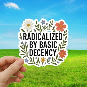 May include: A white sticker with the text "RADICALIZED BY BASIC DECENCY" in black, surrounded by a floral border with white, orange, blue, and yellow flowers and green leaves. The sticker is held against a backdrop of a green field and blue sky.