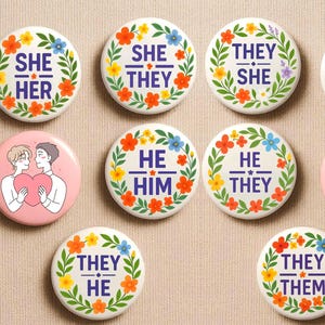 Puede incluir: Una colección de botones redondos con diseños florales y pronombres. Los botones presentan frases como "She/Her", "He/Him" y "They/Them" en una fuente en negrita. Un botón muestra una ilustración de dos personas sosteniendo un corazón. Los botones son blancos con bordes florales coloridos.