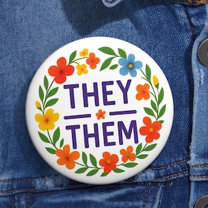May include: A white badge featuring the text "THEY THEM" in purple, encircled by a floral design with orange, yellow, and blue flowers. The badge is set against a denim jacket.