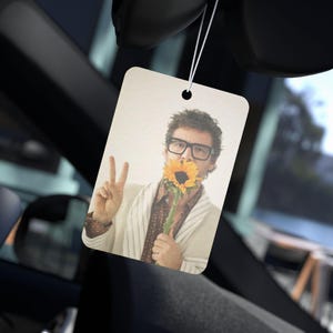 以下が含まれることがあります： ひまわりを持ち、ピースサインをしている人の写真が写った長方形の車の芳香剤。その人はメガネ、柄物のシャツ、ストライプのジャケットを着ています。芳香剤は紐で吊り下げられています。