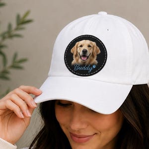 May include: White baseball cap with a circular patch displaying a golden retriever's portrait and the name "Buddy" in blue script. The cap is made of a soft material and has a curved peak. A classic accessory for dog lovers.