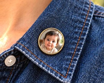 May include: A circular pin-back button featuring a photographic portrait of a young child with dark hair. The button has a metallic gold-toned rim and is attached to the collar of a dark blue denim shirt with visible white stitching and buttons.