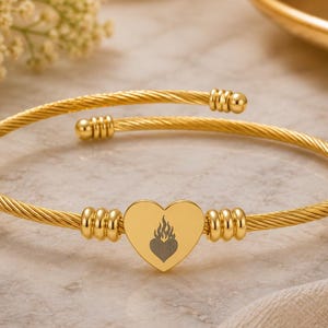 Op de afbeelding: Goudkleurige, open armband. De armband heeft een gedraaid kabelontwerp en een hartvormige bedel met een vlammensymbool. De armband wordt gepresenteerd op een marmeren oppervlak met bloemen en een gouden schaal op de achtergrond.