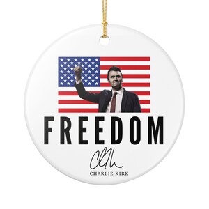 May include: A round ceramic ornament featuring a patriotic theme. The ornament displays an image of a person in a suit with their fist raised in front of an American flag. The word 'FREEDOM' is printed in large, bold, black letters. Below the word is a signature that reads 'Charlie Kirk'.