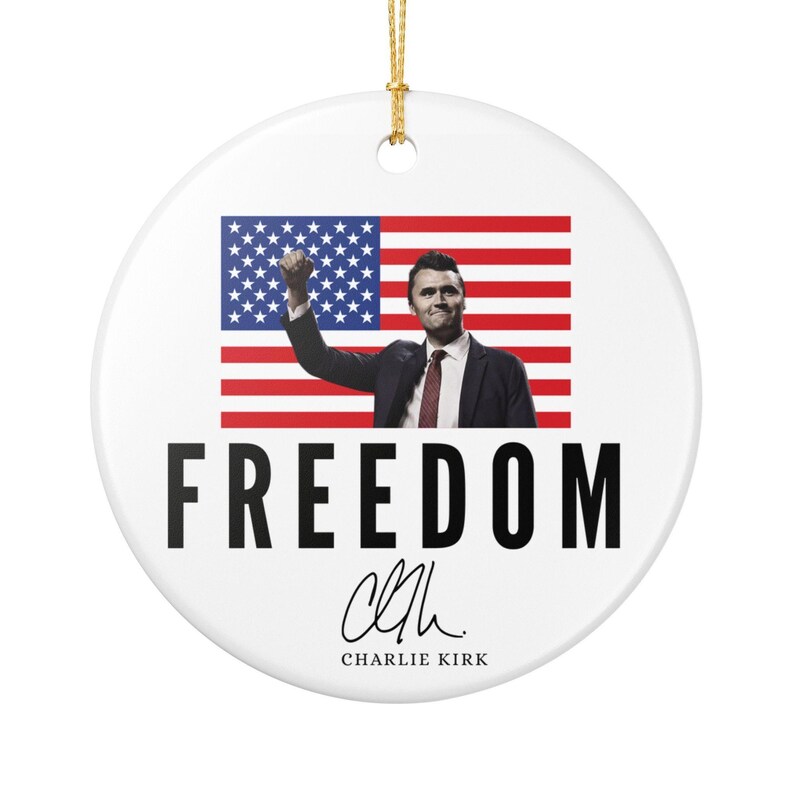 May include: A round ceramic ornament featuring a patriotic theme. The ornament displays an image of a person in a suit with their fist raised in front of an American flag. The word 'FREEDOM' is printed in large, bold, black letters. Below the word is a signature that reads 'Charlie Kirk'.