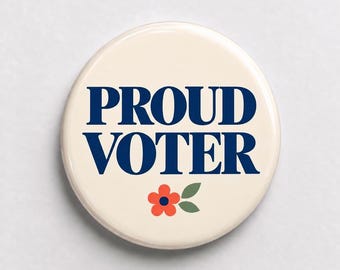 May include: A round pinback button with a cream-colored background. The text PROUD VOTER is printed in bold, dark blue capital letters. Below the text, there is a small graphic of a red flower with a dark blue center and two green leaves.
