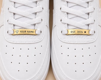 May include: A pair of white sneakers featuring gold-toned metal lace tags. The left tag displays a heart symbol and the text YOUR NAME, while the right tag displays the text EST. 2026 and a star symbol. The shoes have white laces and perforated toes.