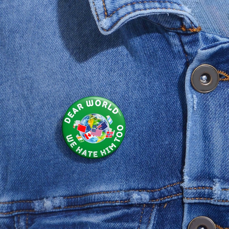 May include: A round green pinback button featuring a central graphic of the Earth surrounded by various national flags. The white text printed around the perimeter of the button reads, "DEAR WORLD WE HATE HIM TOO." The button is attached to blue denim fabric.