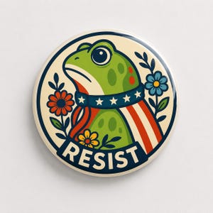 Op de afbeelding: Een ronde button met een groene kikker die een donkerblauwe halsband met witte sterren en een rood-wit gestreepte cape draagt. Het ontwerp bevat rode, blauwe en gele bloemen op een crèmekleurige achtergrond met het woord RESIST in dikke letters.