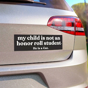 Puede incluir: Una pegatina rectangular negra con texto blanco que dice: "my child is not an honor roll student. He is a Cat." La pegatina est&aacute; aplicada en el maletero plateado de un veh&iacute;culo, colocada horizontalmente debajo de la ventana trasera.