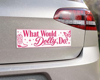 May include: A rectangular bumper sticker with a white background and pink text that reads What Would Dolly Do? The design features a pink western cowboy boot on the left side, a pink butterfly on the right side, and small pink star accents.