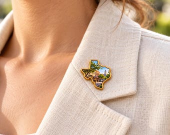 May include: A gold-toned metal pin shaped like the state of Texas. The center of the pin features a detailed, full-color photographic print of a town square with brick buildings, green trees, and a tall clock tower under a bright blue sky.
