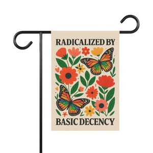 Op de afbeelding: Een tuinvaandel met een crèmekleurige achtergrond met kleurrijke bloemen en twee vlinders met regenboogkleurige vleugels. De tekst "RADICALIZED BY BASIC DECENCY" is op de vlag gedrukt. De vlag is bevestigd aan een zwarte metalen standaard.