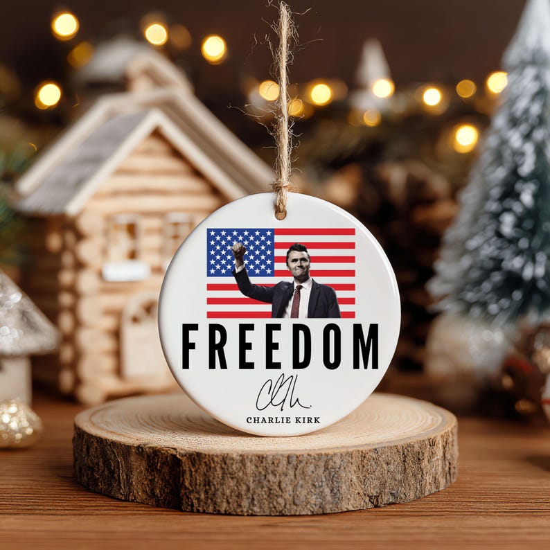 May include: A white ceramic Christmas ornament with a patriotic theme. It features an image of a person in front of an American flag, with the word "FREEDOM" in bold black letters. The ornament is hanging by a natural twine.