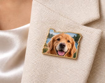 May include: A gold-rimmed pin shaped like the state of Oregon featuring a photographic print of a golden retriever dog with its tongue out, set against a blurred outdoor background of trees and water, attached to a beige textured fabric.