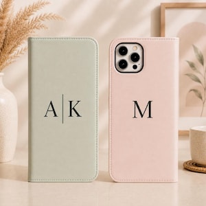May include: A sage green rectangular phone case with the letters A and K separated by a vertical line, and a pale pink phone case with a camera cutout and the letter M. Both cases feature a smooth, matte finish and minimalist typography.
