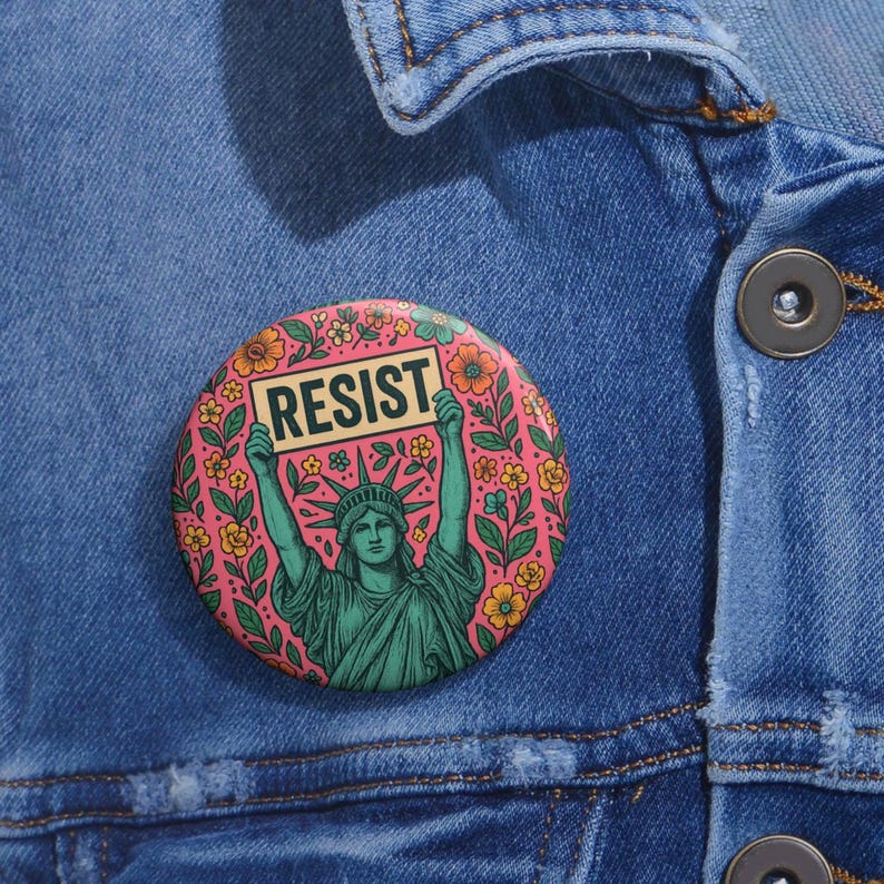 Op de afbeelding: Een roze knoop met een bloemenontwerp met een groen beeld van het Vrijheidsbeeld dat een bord vasthoudt met de tekst "RESIST".