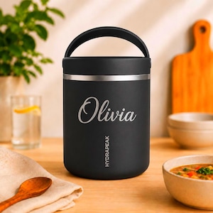 Könnte beinhalten: Ein mattschwarzer, zylindrischer Lebensmittelbehälter mit einem Tragegriff und einem silbernen Rand. Der Name Olivia ist in weißer Schreibschrift auf der Vorderseite eingraviert, darunter steht der Markenname HYDRAPEAK in weißen Großbuchstaben.