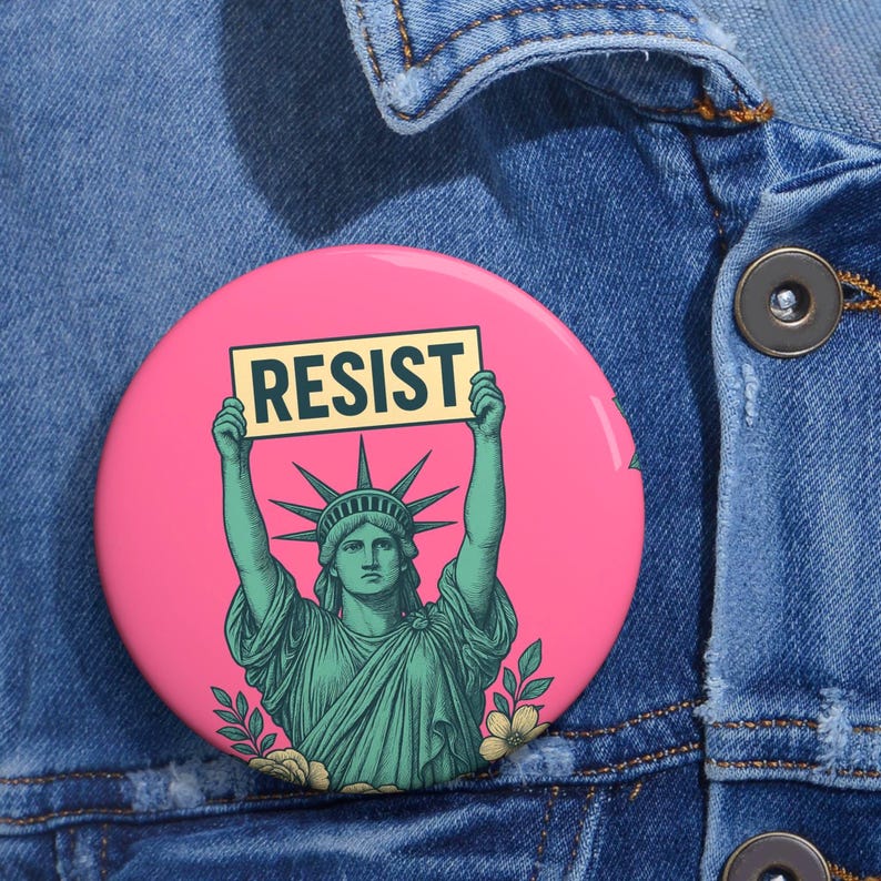May include: A pink button with a green statue of liberty holding a sign that says 'RESIST'.
