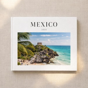 May include: A white hardcover book with the word "MEXICO" and the year "2025" printed on the cover. The book features a vibrant photograph of a coastal scene with turquoise water, a rocky cliff, and a historic structure.