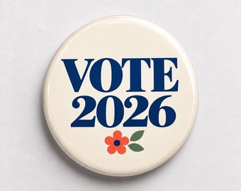 May include: A circular pinback button with a cream-colored background. The text VOTE 2026 is printed in a bold, dark blue serif font. Below the text, there is a small, stylized orange flower with a dark blue center and two green leaves.