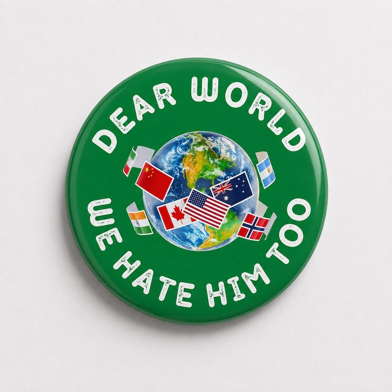 May include: A circular green pin-back button featuring a central image of the Earth surrounded by various national flags. The white text printed in a circular arc around the graphic reads, "DEAR WORLD WE HATE HIM TOO" in a distressed, bold font.