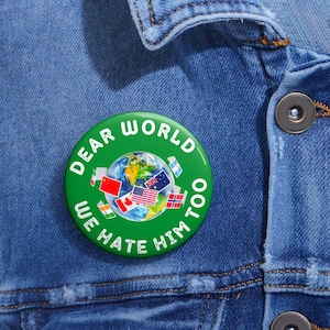 May include: A round green pinback button featuring a central graphic of the Earth surrounded by various national flags. The text "DEAR WORLD WE HATE HIM TOO" is printed in white capital letters in a circular arc around the central globe image.