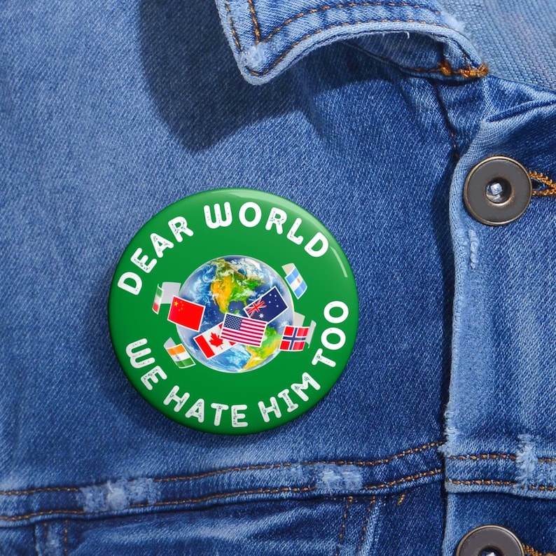 May include: A round green pinback button featuring a central graphic of the Earth surrounded by various national flags. The text "DEAR WORLD WE HATE HIM TOO" is printed in white capital letters in a circular arc around the central globe image.