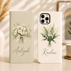 Könnte beinhalten: Zwei rechteckige Handyhüllen im Brieftaschen-Stil. Die linke Hülle ist salbeigrün mit einem Gänseblümchenstrauß und dem Namen Aaliyah. Die rechte Hülle ist cremefarben mit einem Maiglöckchenstrauß, Kameraaussparung und dem Namen Kaelan in Schreibschrift.