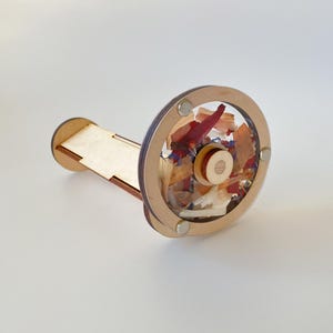 Può includere: Un caleidoscopio in legno con un'area di visualizzazione circolare trasparente riempita di fiori secchi nei toni del rosso, arancione e blu. Ha un manico rettangolare in legno e una cornice circolare in legno.