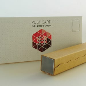Könnte beinhalten: Eine weiße Postkarte mit der Aufschrift "POST CARD kaleidoscope" und einem geometrischen Design in Rot und Braun. Daneben eine braune rechteckige Schachtel mit silbernem Ende.