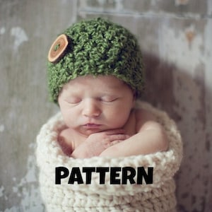May include: A sleeping baby wearing a green crocheted hat with a wooden button. The baby is wrapped in a white crocheted blanket.