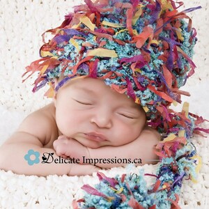 Könnte beinhalten: Ein Baby trägt eine bunte gehäkelte Mütze mit einem langen, flauschigen Schwanz. Die Mütze ist aus verschiedenen Farben gefertigt, darunter Blau, Rot, Gelb und Lila. Das Baby schläft friedlich auf einer weißen Decke.