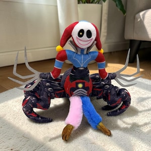 Puede incluir: Un peluche con una figura de bufón con un sombrero rojo y blanco, un torso azul y rojo y una cara sonriente. El juguete tiene múltiples brazos y piernas, con un cuerpo oscuro estampado y piernas multicolores.