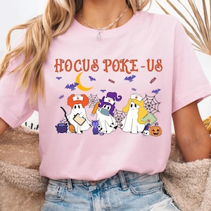 May include: A light pink t-shirt with the text "Hocus Poke-Us" in orange. The shirt features a Halloween-themed graphic of three ghost characters dressed as nurses, with bats, a moon, and spiderwebs.
